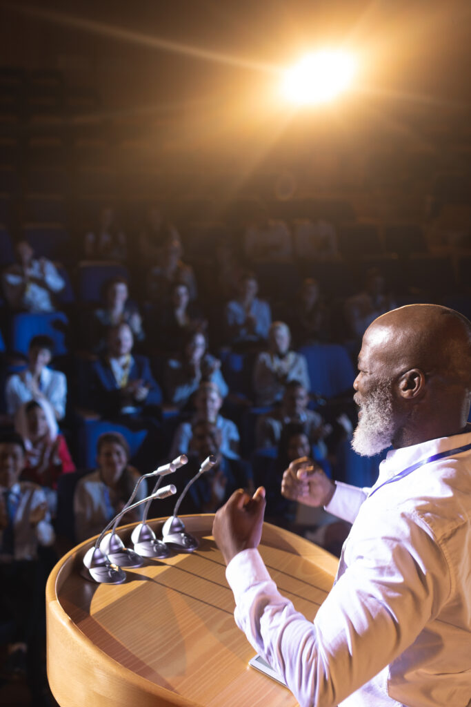 High angle view of old African-American businessman standing near podium and giving speech to the audience in the auditorium